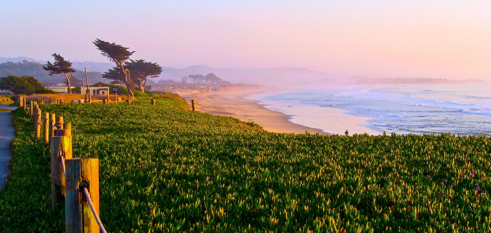 A seaside cliffside path with green coastal plants, wooden posts and rope railing, sunlit beach, gentle waves, and distant hazy hills at sunset.