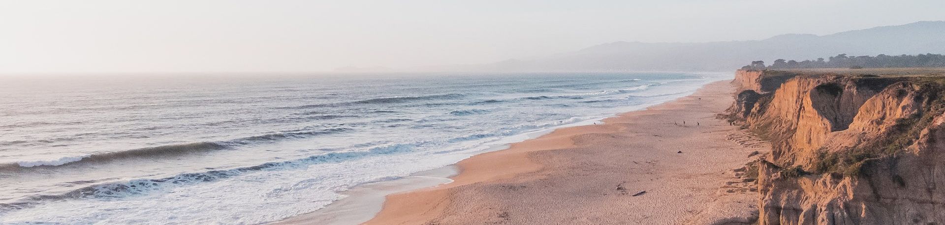 A long sandy beach with gentle waves, cliffs on the right, and distant mountains hazily visible under a soft, pale sky.