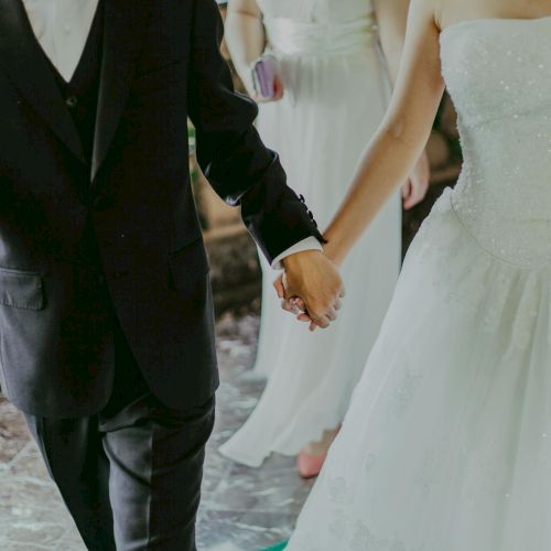 A couple in formal wedding attire holding hands: a man in a black suit and tie, and a woman in a white wedding dress, walking together.