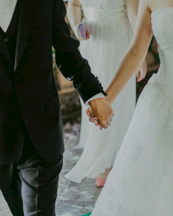 A couple in formal wedding attire holding hands: a man in a black suit and tie, and a woman in a white wedding dress, walking together.