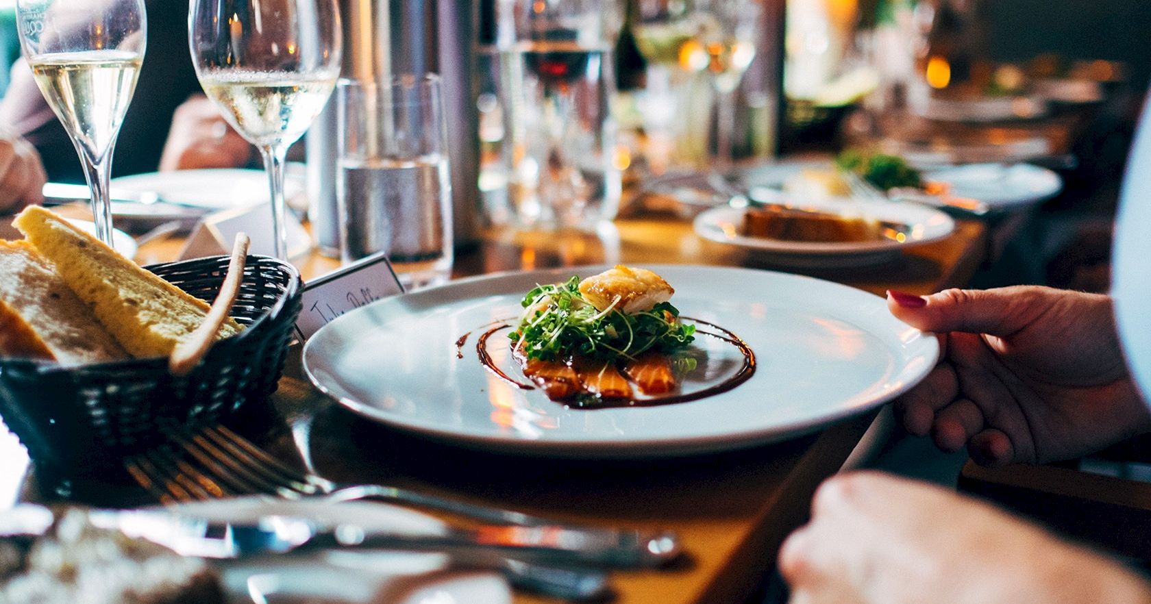A fine dining table scene with plated appetizer, wine glasses, bread basket, and hands readying utensils for a meal.