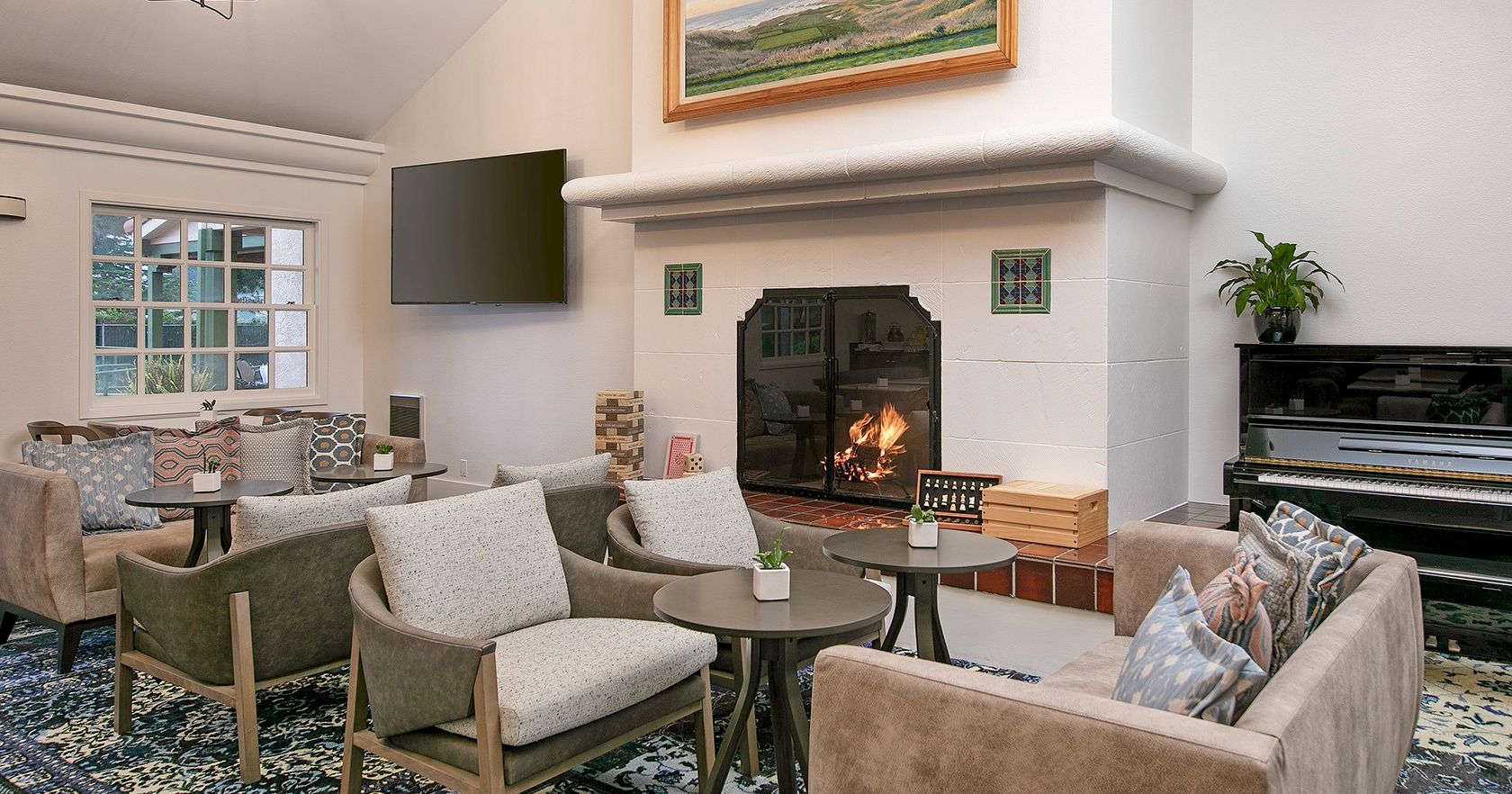 A cozy living room with a lit fireplace, glass coffee table, surrounding beige chairs, a wall-mounted TV, a piano, and a patterned rug, all in soft neutrals.