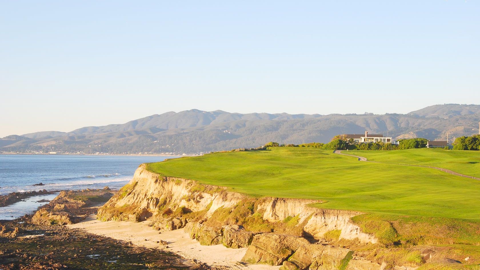 A coastal cliffside with green grassy turf, rocky shoreline, and distant hills by calm blue water, with a few buildings on the horizon.
