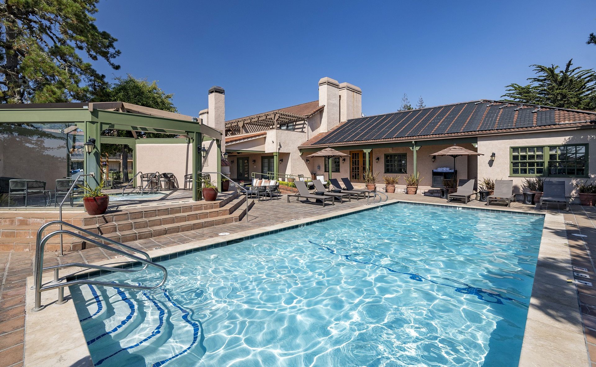 A sunny backyard with a rectangular in-ground pool, blue water, sun loungers, a patio, and a beige house with a tiled roof and solar panels, under a clear sky.