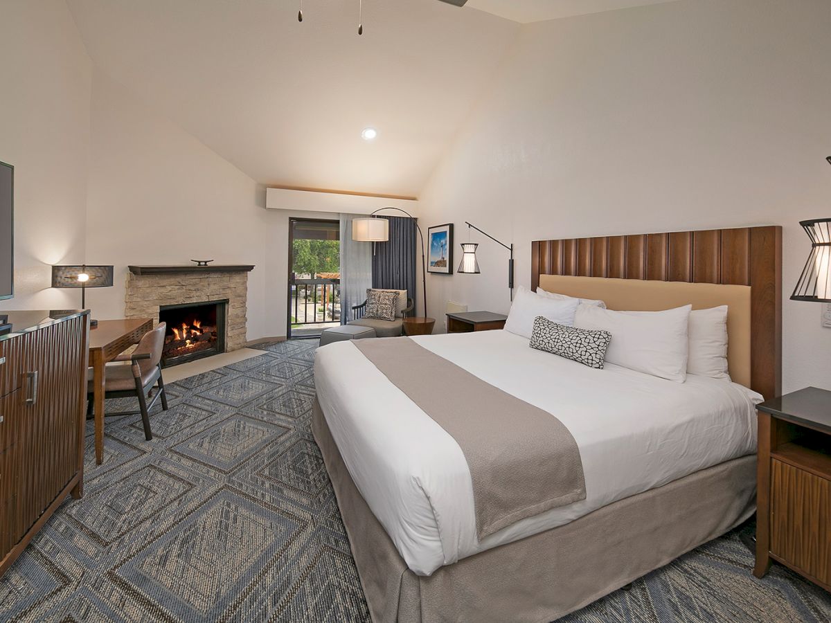 A modern hotel room with a king bed, wooden headboard, seating area, fireplace, flat-screen TV, and a sliding door to a balcony, stylish and cozy.