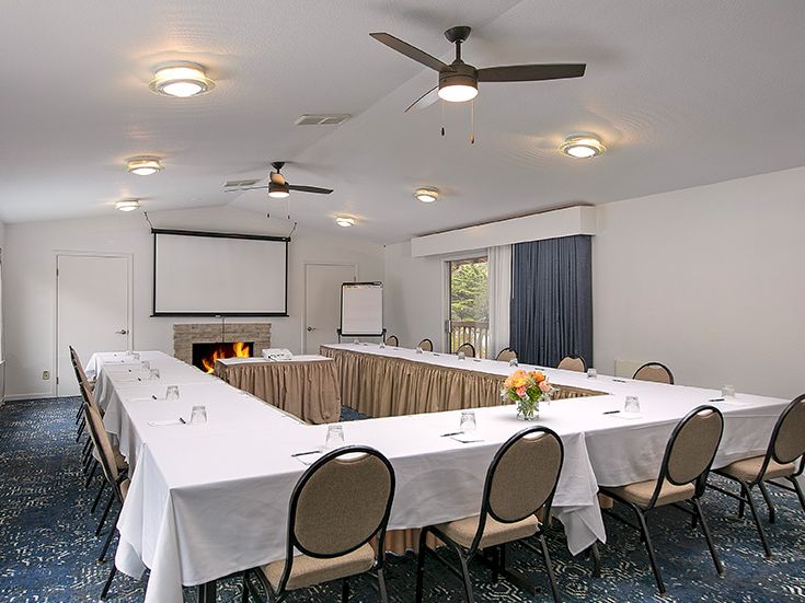 A conference room set for a meeting with U-shaped tables, white tablecloths, chairs, a projector screen, a fireplace, and ceiling fans.