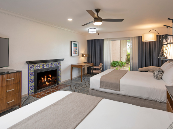 A modern hotel room with two queen beds, a flat-screen TV, a fireplace, a wooden dresser, seating area, desk, and a glass door to a patio, all in warm neutrals.