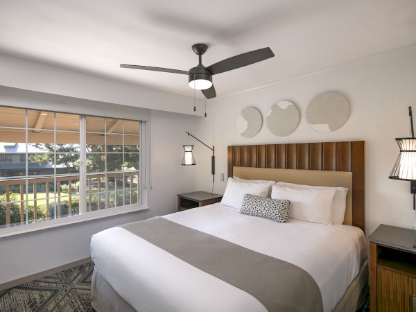 A bright hotel bedroom with a king bed, wood headboard, two nightstands, modern pendant lamps, a ceiling fan, large window, and neutral gray bedding, ending with a period.