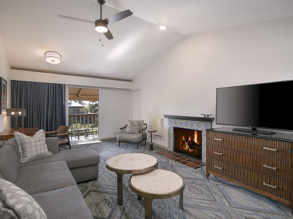 Cozy living room with a gray sectional, round coffee tables, fireplace, large TV on a wooden console, and a balcony with blue curtains in the background.