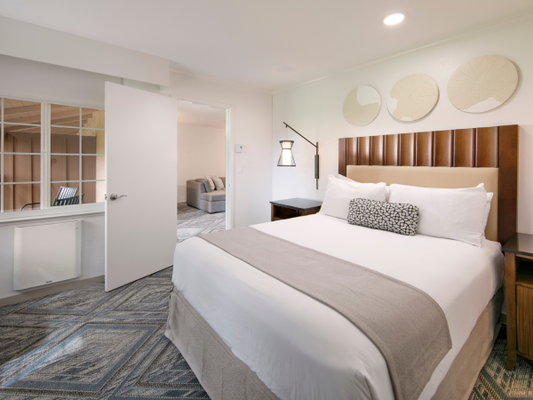 A modern hotel room with a large bed, nightstands, wall sconces, and geometric carpet; ensuite doorway leads to a seating area.