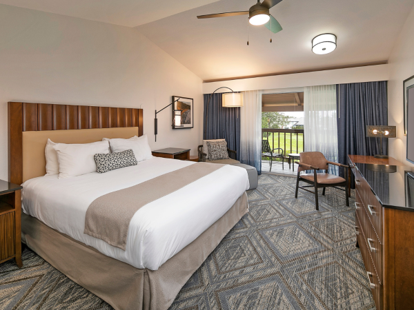 A hotel room with a large bed, nightstands, a desk, a chair, a TV, and a small balcony visible through glass doors, modern decor, neutral tones, and ceiling fan.