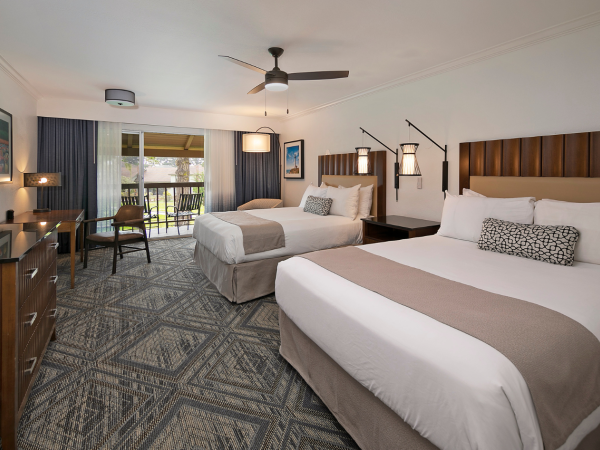 A spacious hotel room with two queen beds, a dark wood headboard, a desk and chair, a balcony view, and blue-gray patterned carpet, bright and modern.