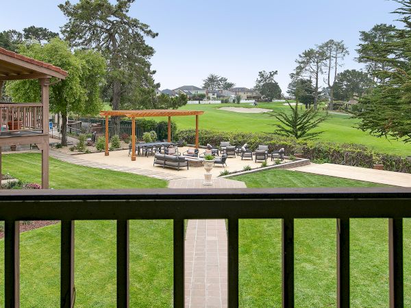 A fenced balcony overlooks a well-kept lawn and outdoor seating area with a pergola, patio, and green fields beyond the trees.