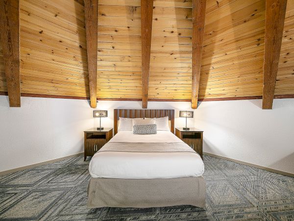 A cozy attic-style bedroom with a centered bed, wooden beams overhead, two nightstands, and soft lighting, creating a warm, cabin-like vibe.