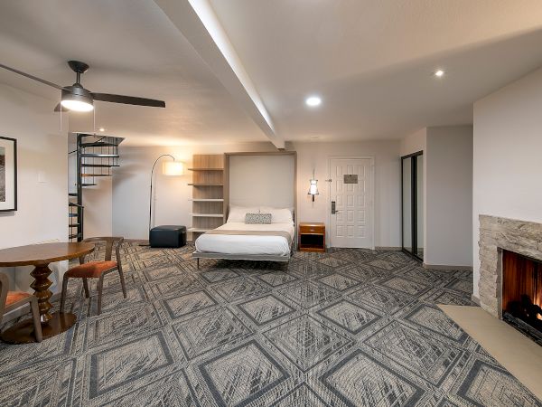 A modern hotel room with a bed, fireplace, round dining table, spiral staircase, and a cozy seating area under soft lighting. The text ends here.