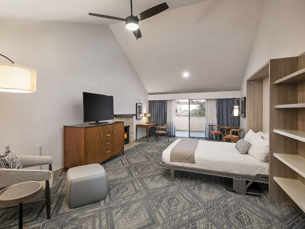 A spacious hotel suite with a king bed, a seating area, a TV on a dresser, a desk, and open shelving in warm neutral tones. End.