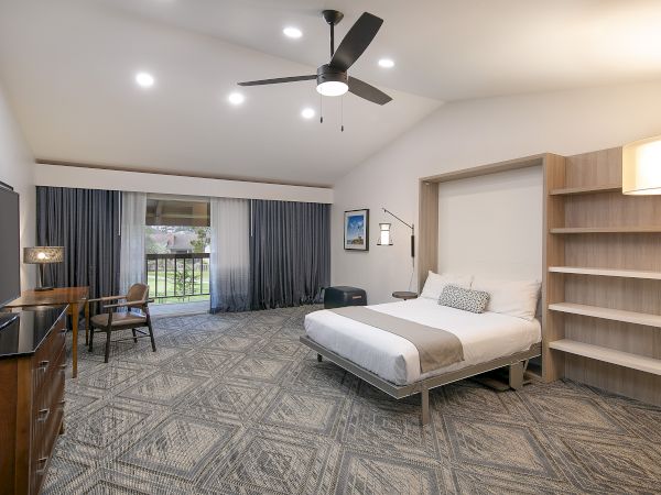 A modern hotel room with a king bed, built-in shelves, a desk and chair, a TV, ceiling fan, and large sliding glass doors with dark curtains.