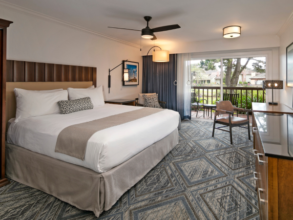 A spacious hotel room with a king bed, wooden armoire, desk, two chairs, and a balcony view; neutral tones and patterned carpet, ceiling fan.