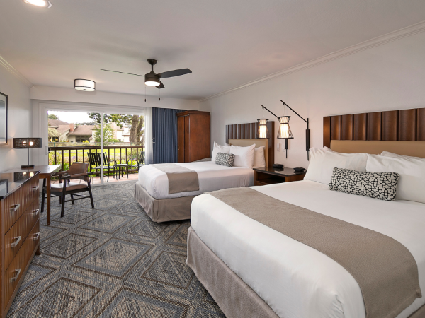 A bright hotel room with two queen beds, a desk, flat-screen TV, and a small seating area, opening to a balcony overlooking a landscaped courtyard.