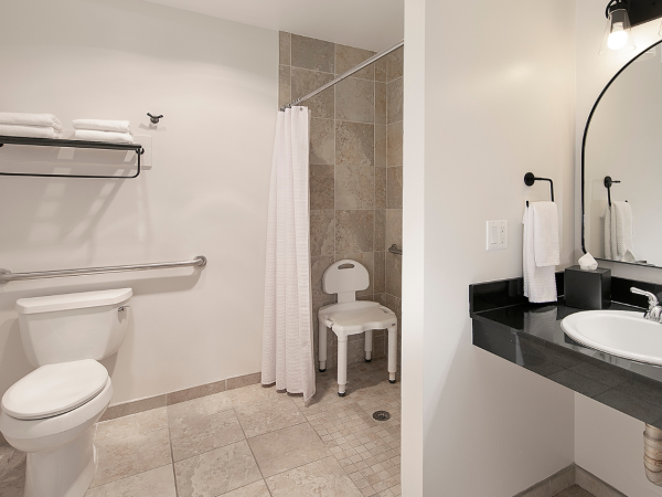 A clean accessible bathroom with a toilet, grab bars, shower, white curtains, and a black granite sink counter with a round mirror, ending with a period.