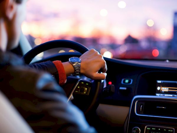 Someone driving a car at dusk, hands on the steering wheel, dashboard lights glow, city lights blur outside the windshield.