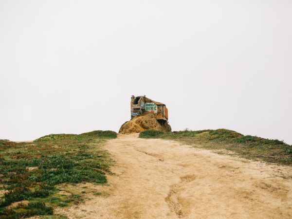 A dirt path leads to a small, weathered house perched on a hilltop, sparse grass on either side under a pale sky.