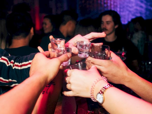 Friends toast with shot glasses in a dim club, raised in a circle over a red-lit bar. Ending with a period.