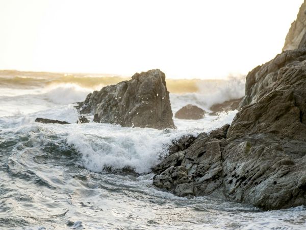 Rugged coastline with jagged rocks and crashing waves, sunlight glints off the water as spray erupts around the rocks.