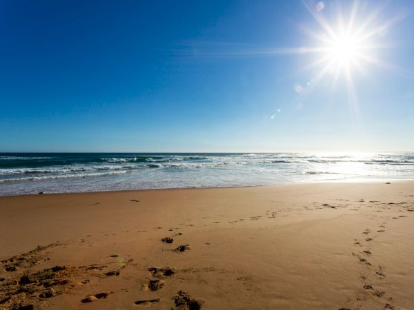 A sunny beach with clear blue sky, sparkling sea, and footprints in the golden sand under a bright sun over calm waves.