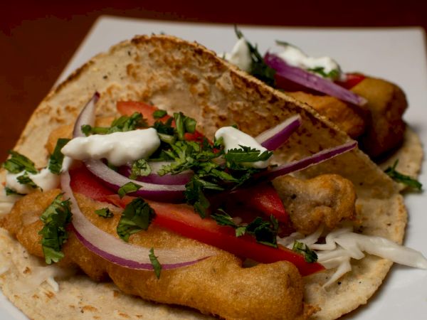 A fish taco on a tortilla topped with onions, cilantro, tomatoes, and a dollop of crema, served on a plate with lime wedges.