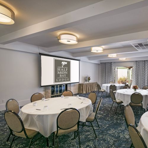 A banquet hall set for a formal event with round tables, white linens, and a projector screen displaying the logo, ready for guests.