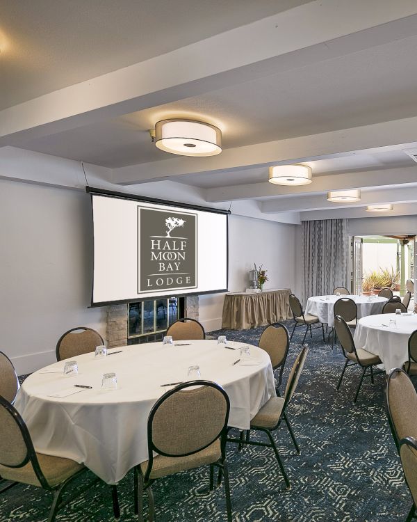 A banquet hall set for a formal event with round tables, white linens, and a projector screen displaying the logo, ready for guests.