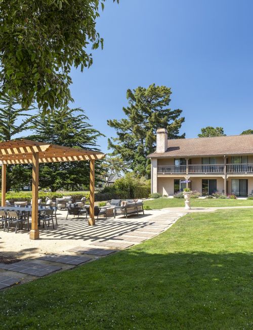 A sunny outdoor garden area with a wooden pergola, patio seating, a well-kept lawn, and a two-story beige building in the background.