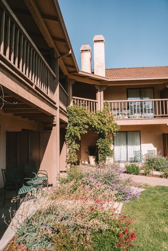 A two-story residential courtyard with balconies, planters, and a sunny lawn, arching entryways, and climbing vines along the wall. End.