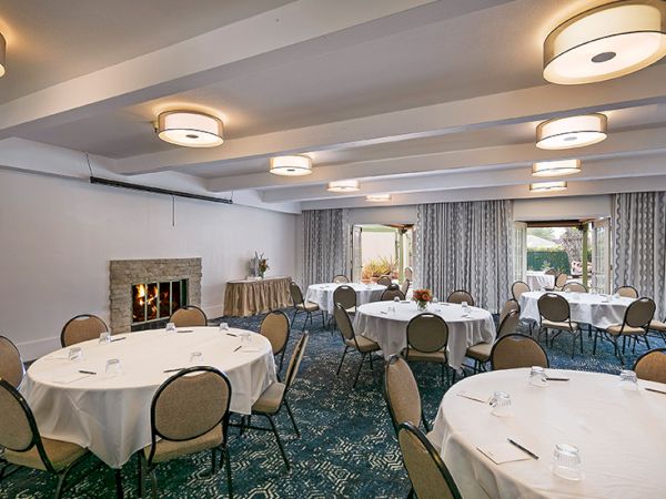 A banquet room set for a function with round tables, white linens, cushioned chairs, and soft overhead lighting in a cozy, carpeted hall.