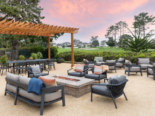 Outdoor modern patio with cushioned seating around a central fire pit, a wooden pergola, gravel ground, and a scenic green landscape under a pink sunset.