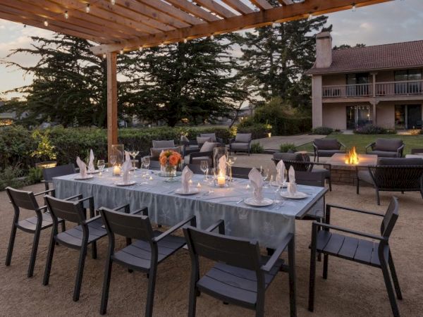 An elegant outdoor dining setup with a long table, candles, folded napkins, and a wooden pergola overlooking a garden and building in the background.