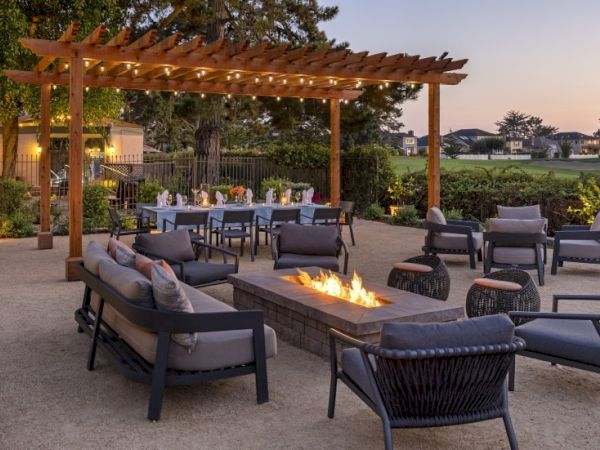 Outdoor lounge area with a wooden pergola, fire pit, and modern seating; a long dining table in the background under sunset skies.