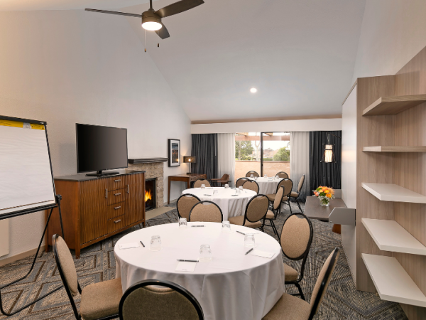 A conference room setup with round tables, cushioned chairs, a fireplace, a flat-screen, a flip chart, and a windowed backdrop for meetings.