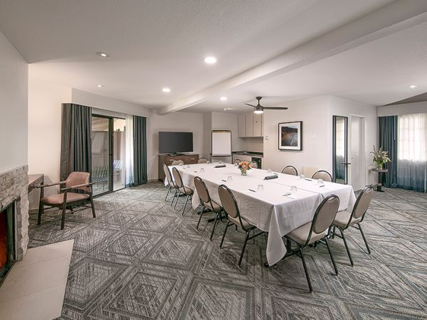 A bright conference room with a long table set for a meeting, beige chairs, a fireplace, and large windows letting in natural light.