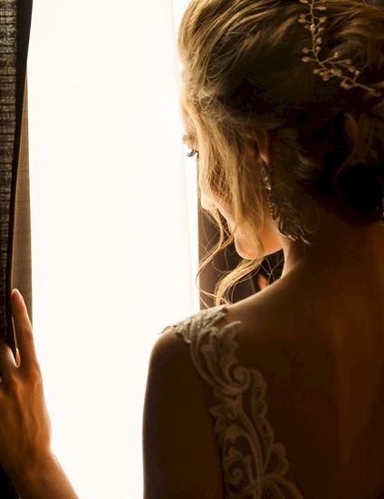 A woman with an elegant updo looks out a window, lace dress back visible, soft backlight enhancing her silhouette.