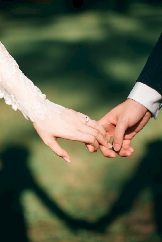 A bride and groom hold hands in a wedding moment, outdoors, fingers entwined, sunlight and shadows on the grass behind.