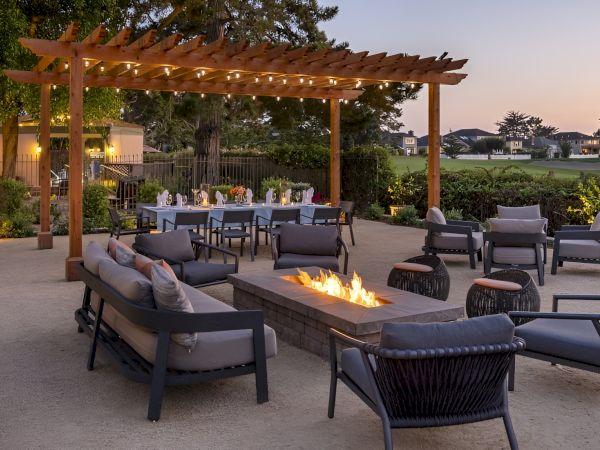 A modern outdoor seating area with a wooden pergola, sofas, chairs, and a rectangular fire pit, set for a gathered evening event.