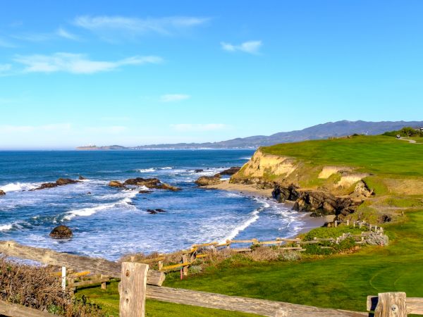 Coastal cliffside with a grassy, fenced foreground, rocky shoreline, blue ocean, and clear sky; a serene seaside landscape.