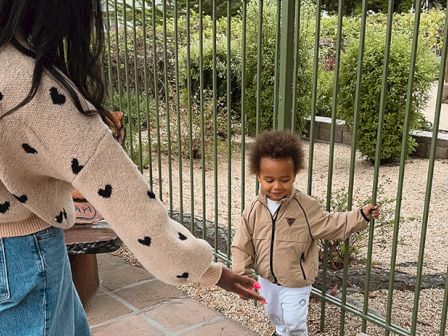 A woman extends a hand to a toddler at a metal gate; they&rsquo;re outdoors near a fenced garden, a joyful moment of connection. End.