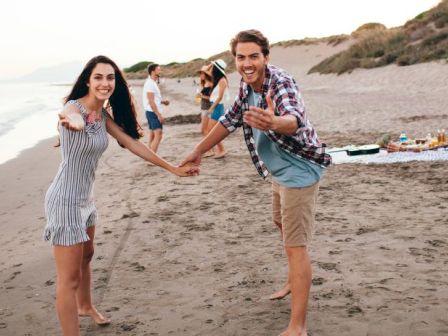 Two friends on a beach hold hands and smile at the camera, with others in the background near a blanket and surf gear.