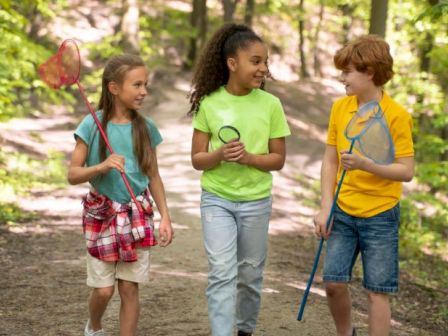 Three kids walk a forest path with nets and a stick, chatting happily as they explore the woods.