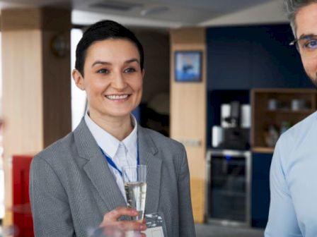 Two professionally dressed people at a social gathering toast with smiles in a modern office setting, holding champagne flutes, and wearing name badges.