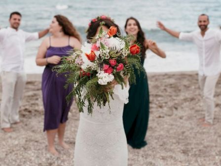 A bride holds a bouquet toward the camera on a beach, with four attendants dancing behind her in colorful outfits, enjoying a joyful moment by the shoreline.