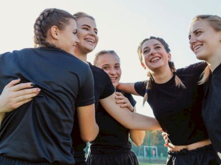 A group of young female athletes in black uniforms huddle together, arms around each other, smiling during a team moment on the field.